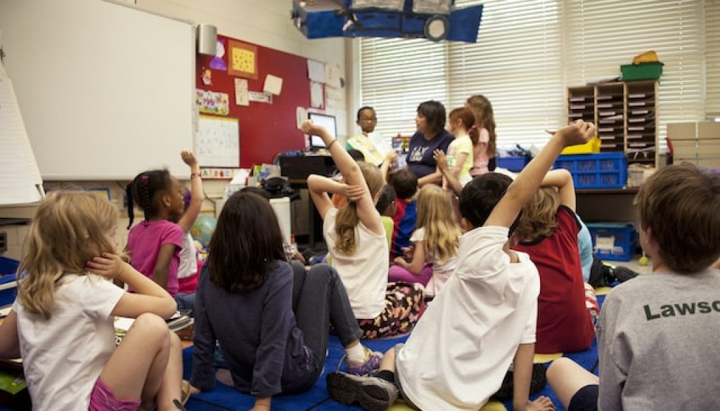 Captured in a metropolitan Atlanta, Georgia primary school, this photograph depicts a typical classroom scene, where an audience of school children were seated on the floor before a teacher at the front of the room, who was reading an illustrated storybook, during one of the scheduled classroom sessions. Assisting the instructor were two female students to her left, and a male student on her right, who was holding up the book, while the seated classmates were raising their hands to answer questions related to the story just read.